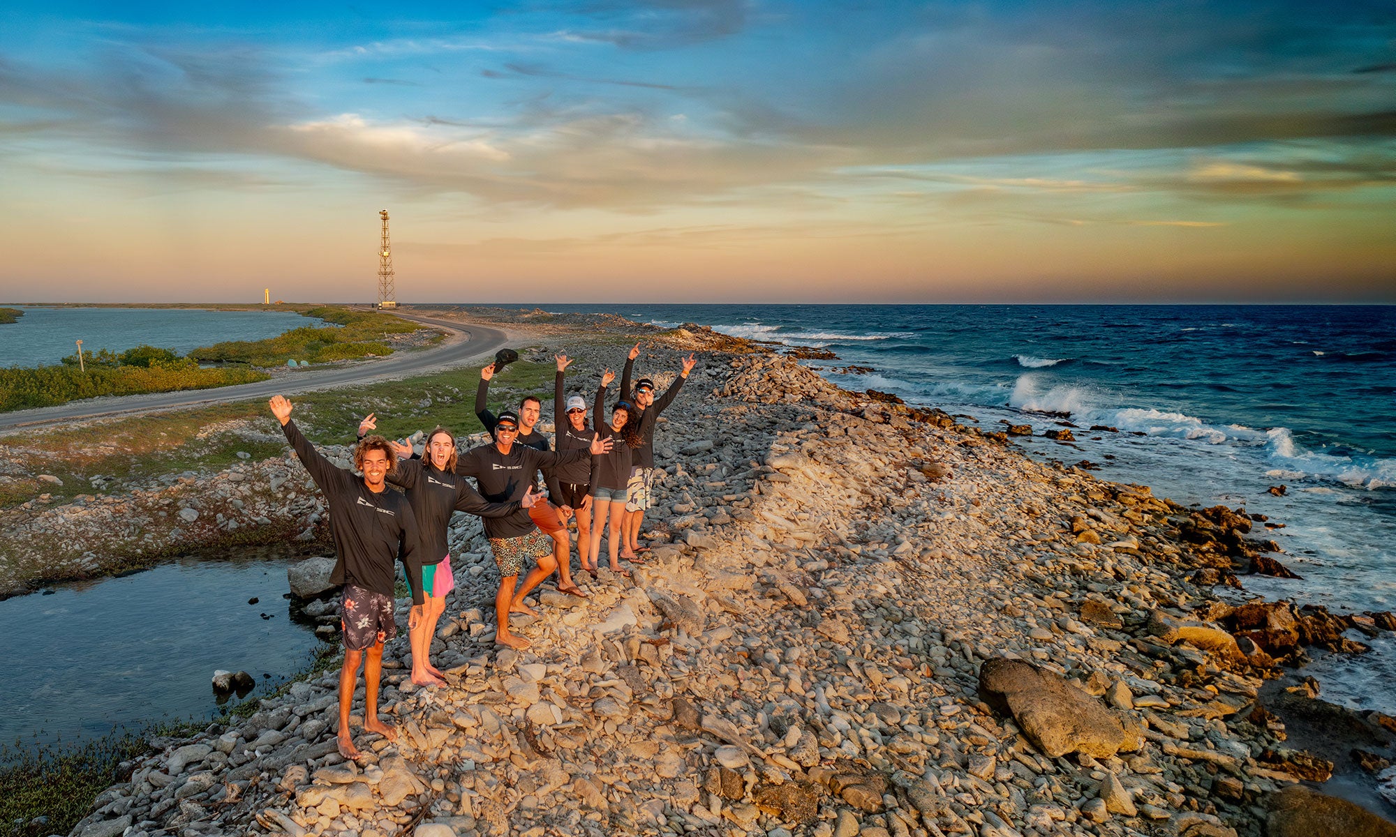 SIC MAUI Athletes Group of people posing on a rocky coastal path with a lighthouse in the distance.