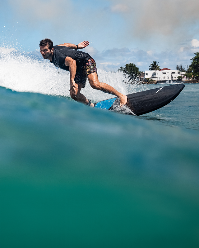 Person surfing on a wave with a building and trees in the background on his Phantom soft surfboard by SICMAUI
