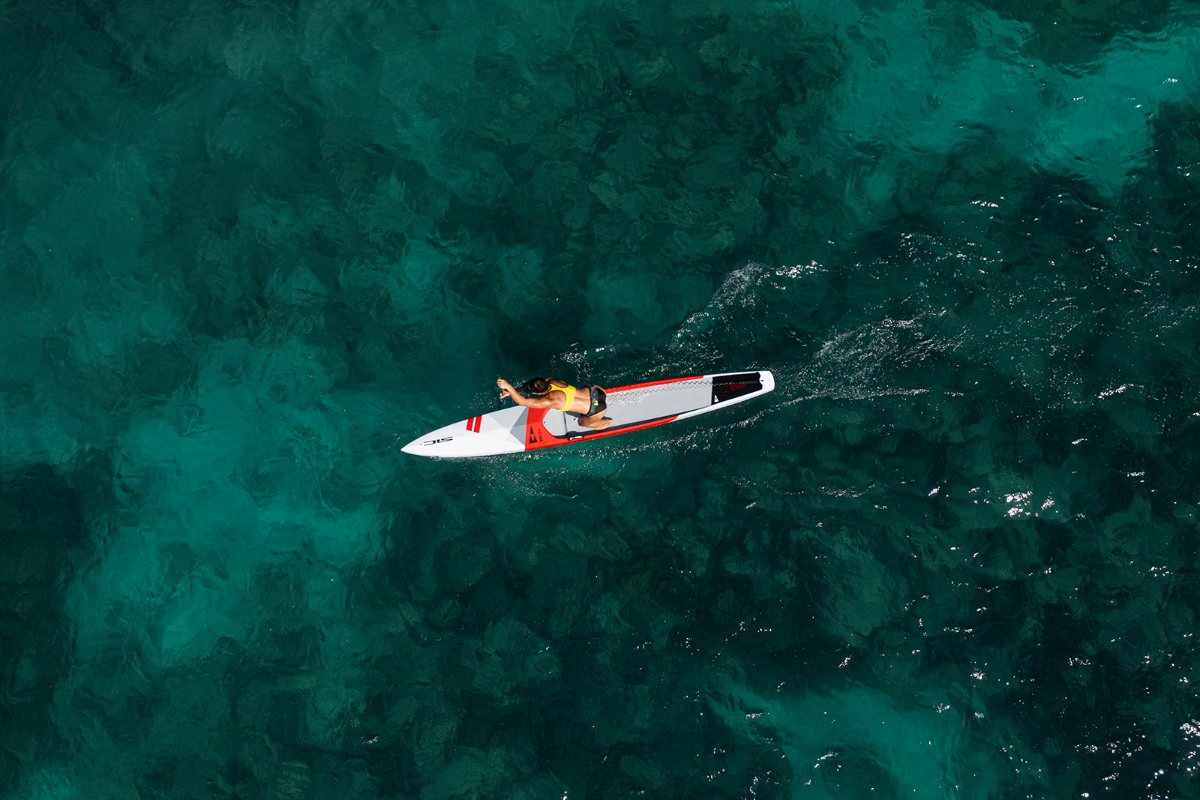 Aerial view of an athlete paddling a SIC Maui race paddleboard in clear turquoise ocean water.