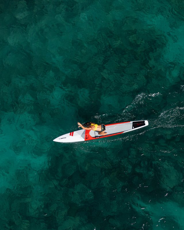Aerial view of an athlete paddling a SIC Maui race paddleboard in clear turquoise ocean water.