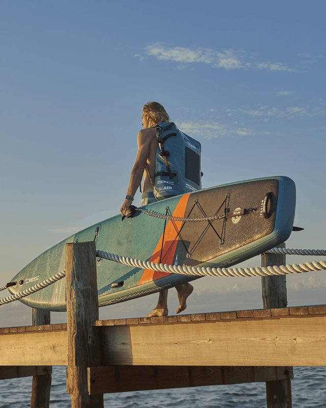 Paddler walking on a pier carrying an inflatable SIC Maui touring paddleboard and a blue SIC Maui dry bag backpack.