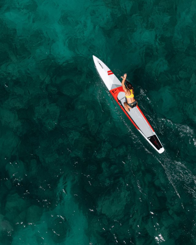 Aerial view of an athlete paddling a SIC Maui race paddleboard in clear turquoise ocean water.