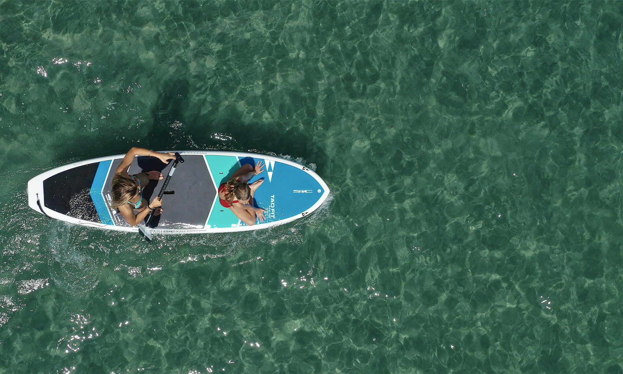 Bird’s-eye shot of a paddleboarding tandem experience on clear emerald ocean waters, showcasing the stability of a SIC Maui board.