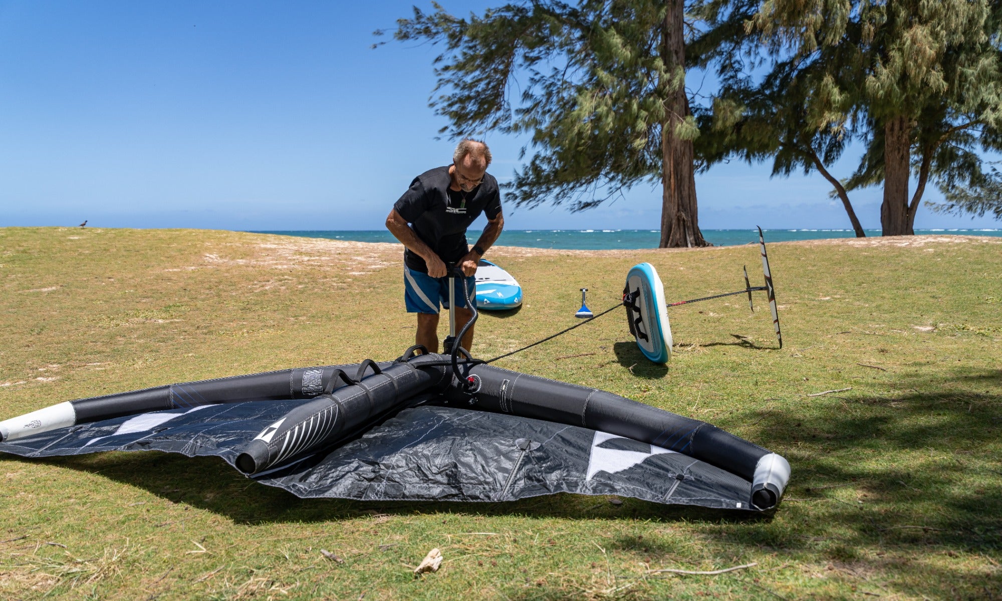 Action shot of inflating a black SIC Maui wing on a tropical grassy beach near the ocean.