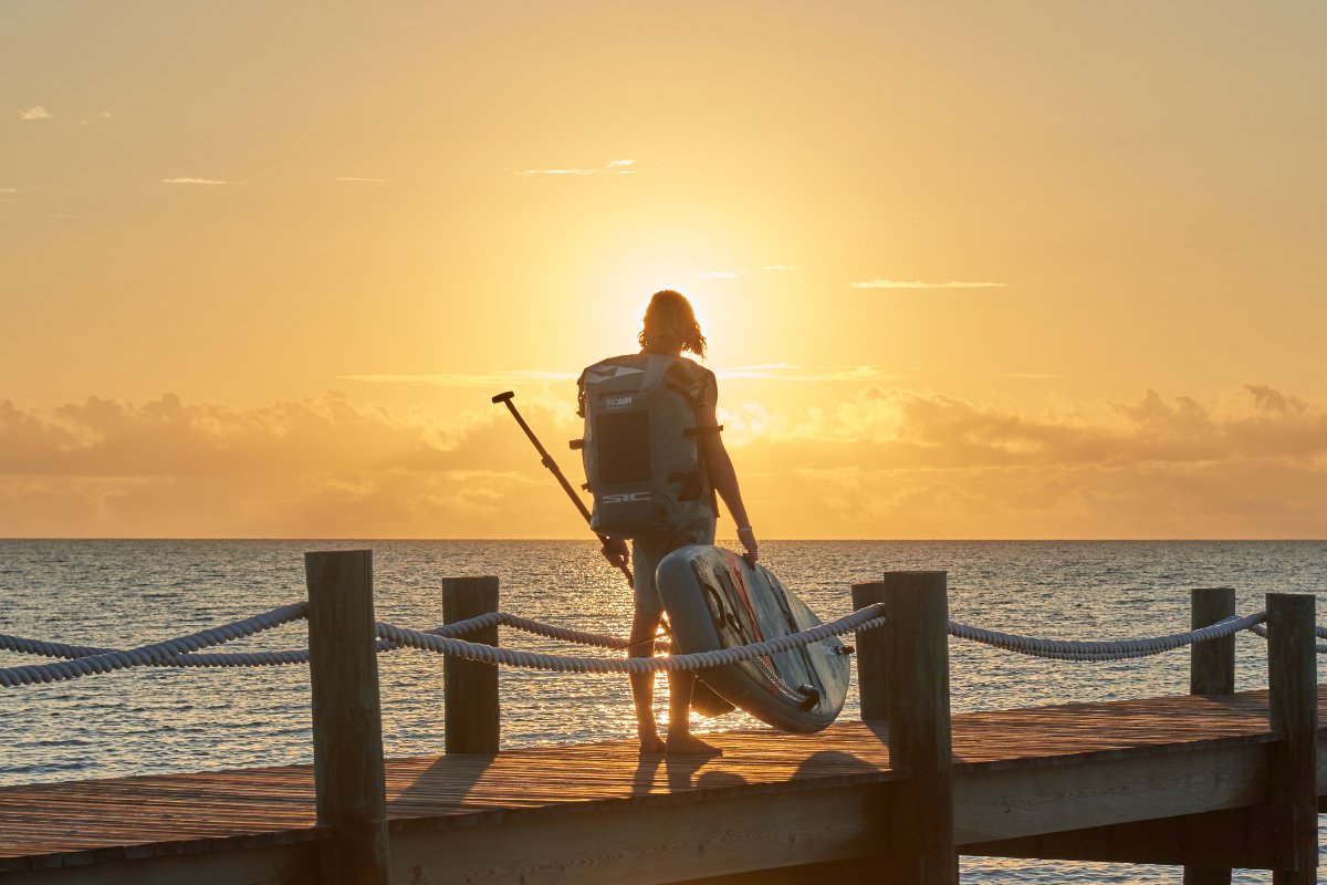 Person with a backpack and paddle standing on a dock at sunset SIC SUP