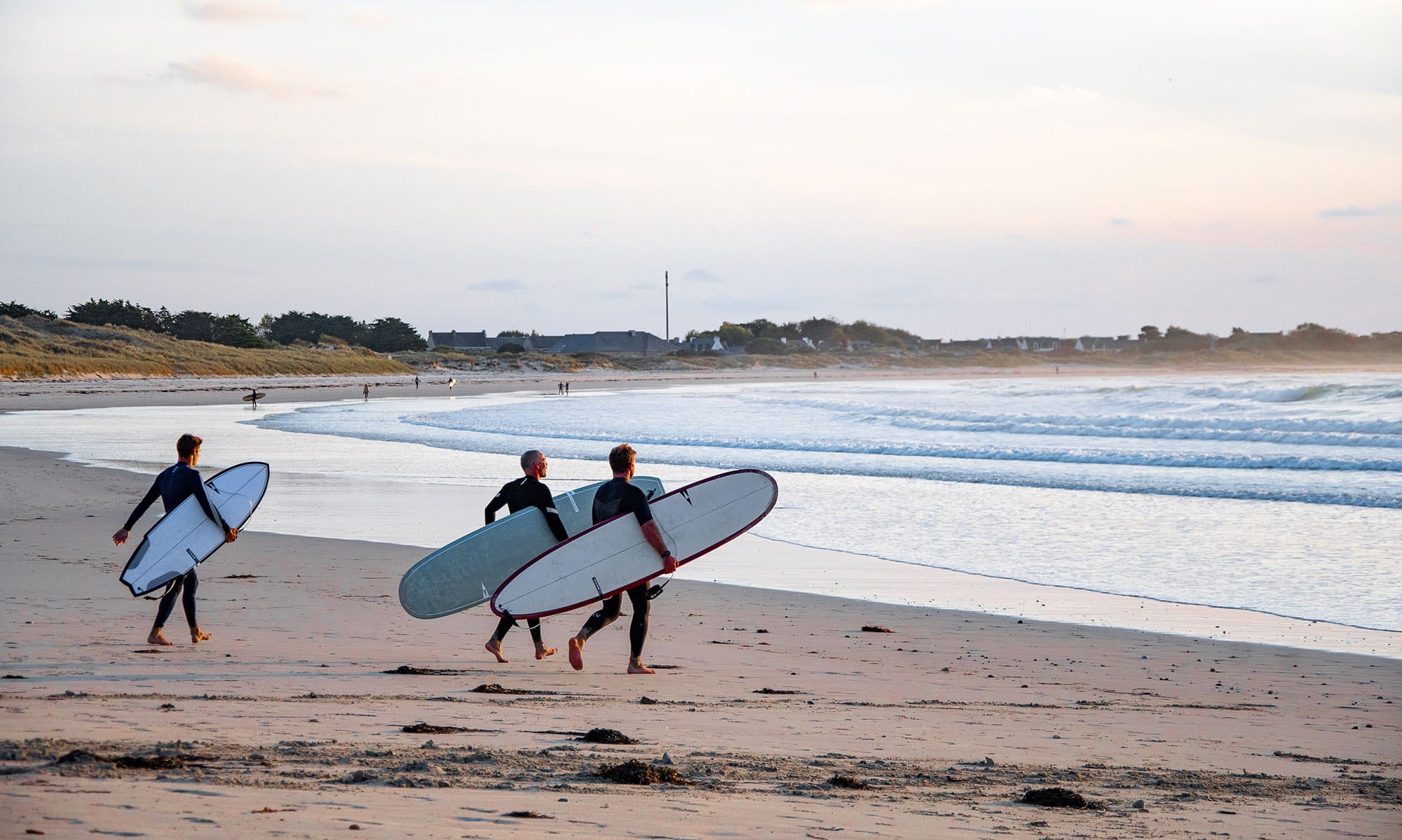 Three surfers walking on a beach with surfboards under a clear sky. with SIC Surfboards composite